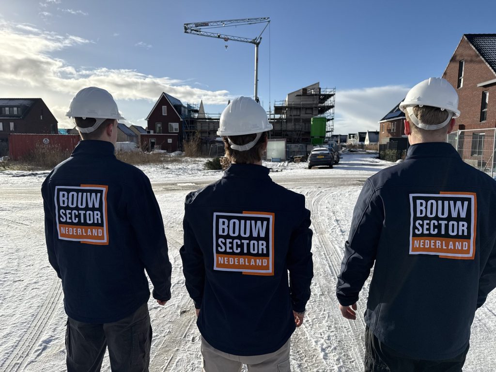 Drie vakmensen van Bouwsector Nederland met witte helmen en bedrijfsjas kijken naar nieuwbouwwoningen met een bouwkraan erachter
